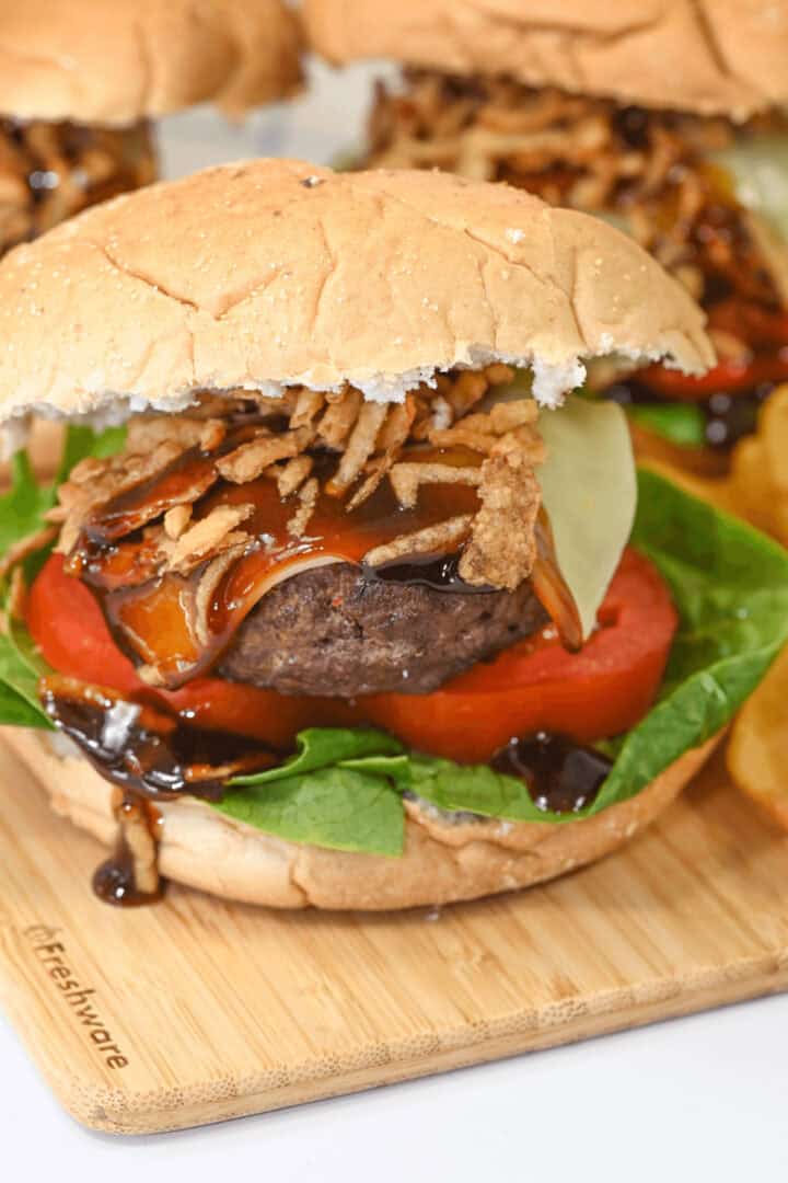A close-up of a Bourbon Burger with lettuce, tomato, cheese, crispy onions, and sauce, served on a wooden board.