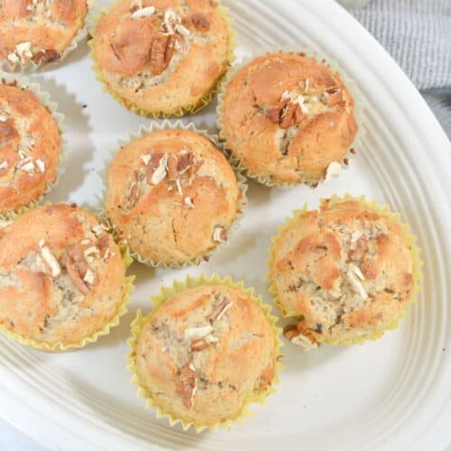 A white platter with nine cinnamon muffins in yellow paper liners, topped with chopped nuts, next to a glass of milk.