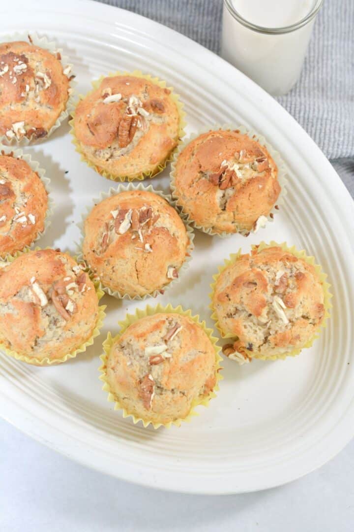 A white platter with nine cinnamon muffins in yellow paper liners, topped with chopped nuts, next to a glass of milk.