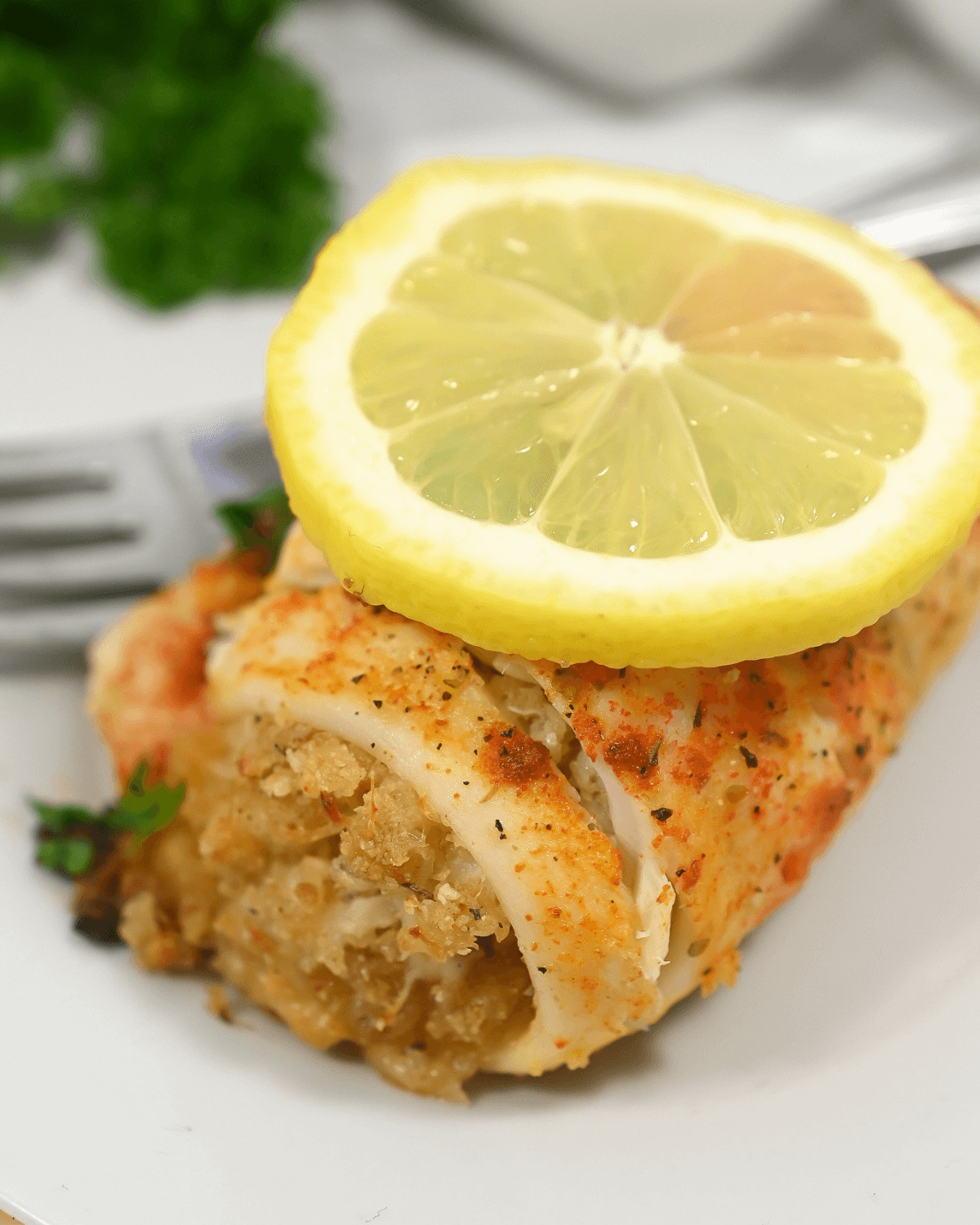 A close-up of a seasoned fish fillet with crabmeat stuffing, garnished with a lemon slice and parsley on a white plate.