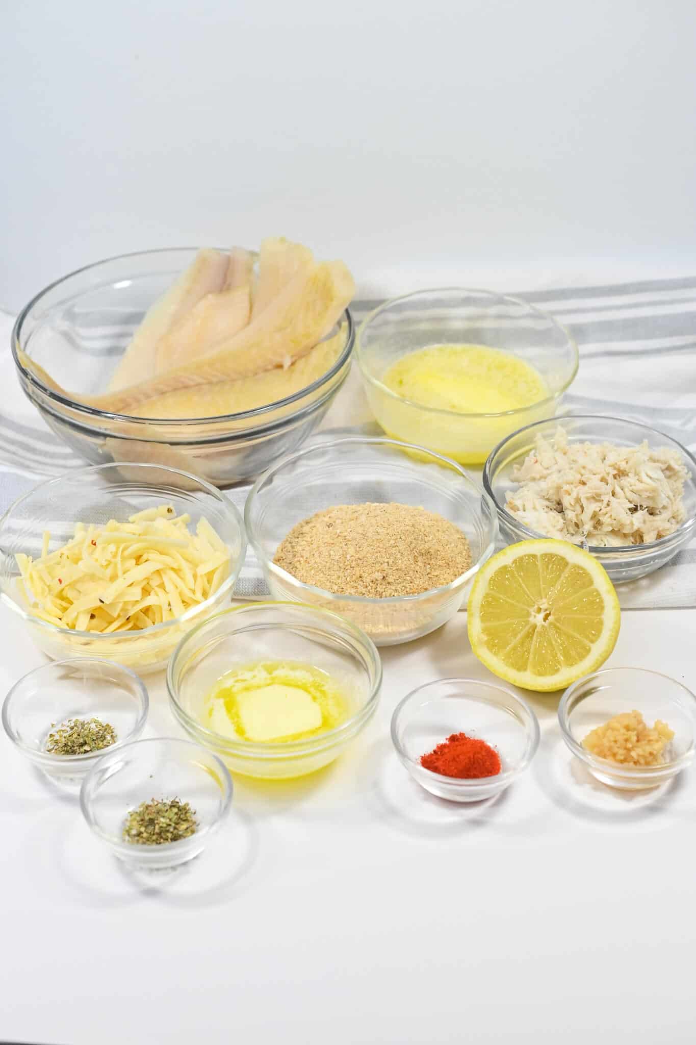 Ingredients for a fish dish laid out on a table, including fish fillets, crabmeat stuffing, breadcrumbs, herbs, spices, and lemon.