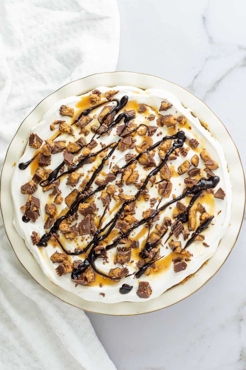 A round Chocolate and Peanut Butter Pie topped with whipped cream, chopped peanut butter cups, caramel sauce, and a chocolate drizzle on a white plate.