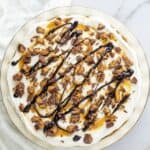 A round Chocolate and Peanut Butter Pie topped with whipped cream, chopped peanut butter cups, caramel sauce, and a chocolate drizzle on a white plate.