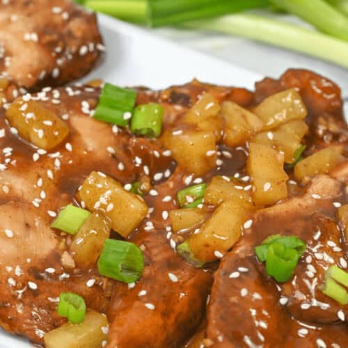 Close-up of slow cooker honey garlic chicken, glazed and garnished with pineapple chunks, sesame seeds, and chopped green onions, served on a white plate with fresh green onions in the background.