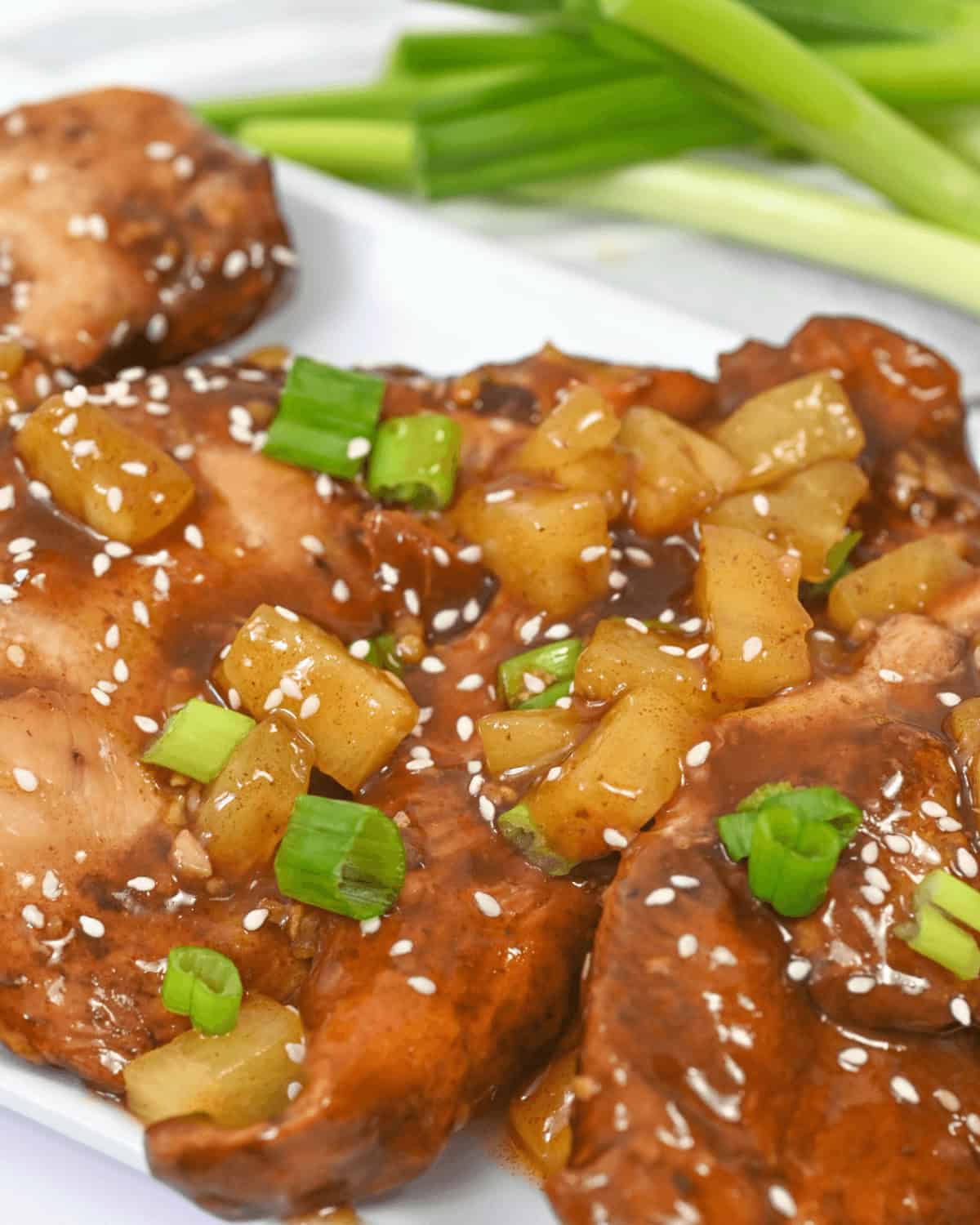 Close-up of slow cooker honey garlic chicken, glazed and garnished with pineapple chunks, sesame seeds, and chopped green onions, served on a white plate with fresh green onions in the background.