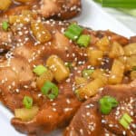 Close-up of Slow Cooker Honey Garlic Chicken glazed pieces garnished with diced pineapple, green onions, and sesame seeds on a white plate, with green onions in the background.
