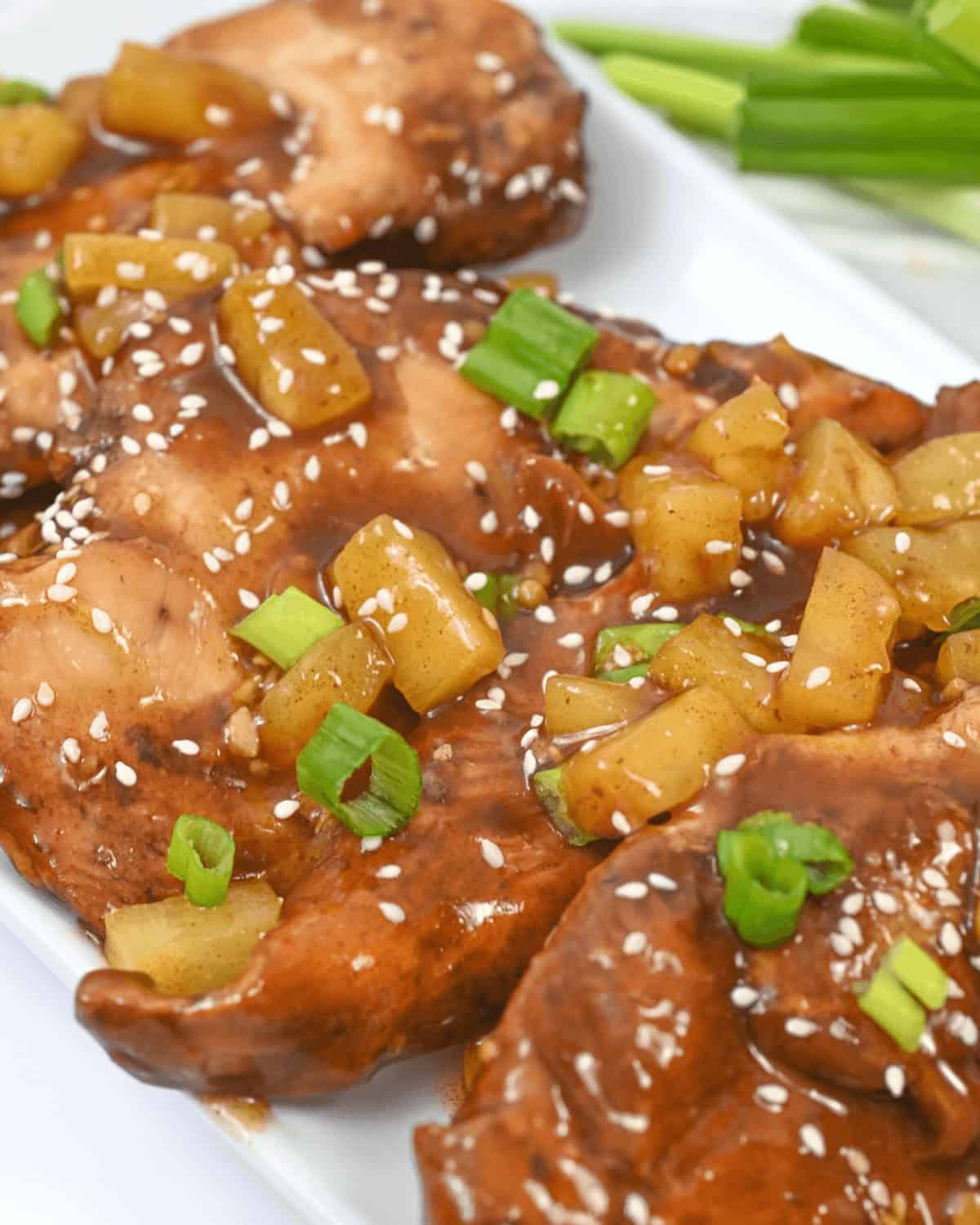 Close-up of Slow Cooker Honey Garlic Chicken glazed pieces garnished with diced pineapple, green onions, and sesame seeds on a white plate, with green onions in the background.