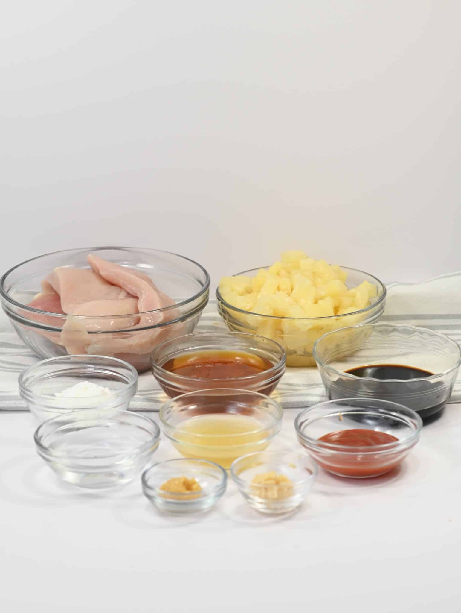 A collection of bowls containing raw chicken breast, pineapple chunks, soy sauce, ketchup, honey, cornstarch, ginger, garlic, water, and pineapple juice sits on a white surface—perfect ingredients for Slow Cooker Honey Garlic Chicken.
