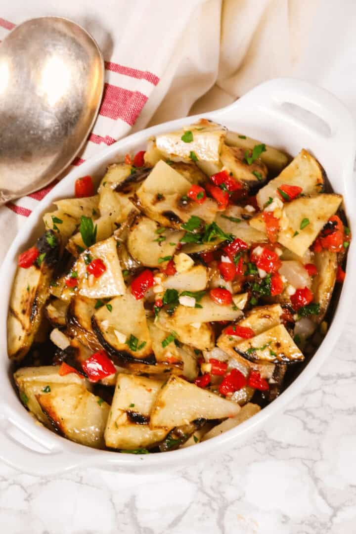A white dish filled with warm potato salad featuring grilled potato wedges, diced red bell peppers, and garnished with chopped herbs. A metal spoon and a folded cloth are next to the dish on a marble surface.