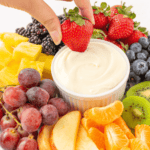 A hand dipping a strawberry into a bowl surrounded by an assortment of fresh fruits.