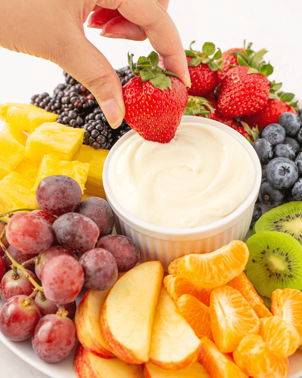 A hand dipping a strawberry into a bowl surrounded by an assortment of fresh fruits.