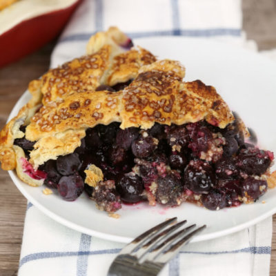 blueberry pie on a white plate on a white checkered napkin with a silver fork.