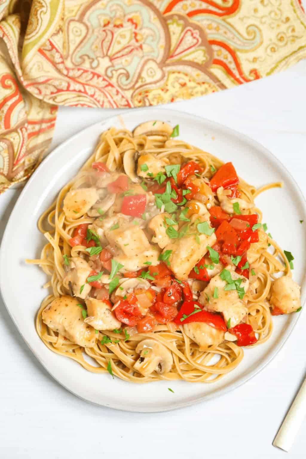 A plate of easy chicken cacciatore: spaghetti topped with seasoned chicken pieces, diced tomatoes, and herbs, placed beside a patterned cloth napkin.