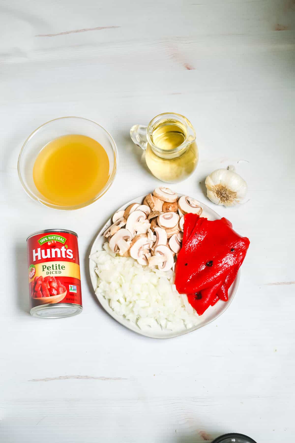 Ingredients for the dish: canned tomatoes, sliced mushrooms, diced onions, sliced red peppers, a whole garlic bulb, a pitcher of broth, and a small jug of oil adorn the table.