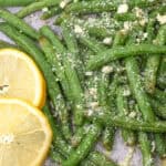 Garlic Parmesan green beans topped with grated cheese sit on a baking sheet, accompanied by two lemon slices for a zesty finish.