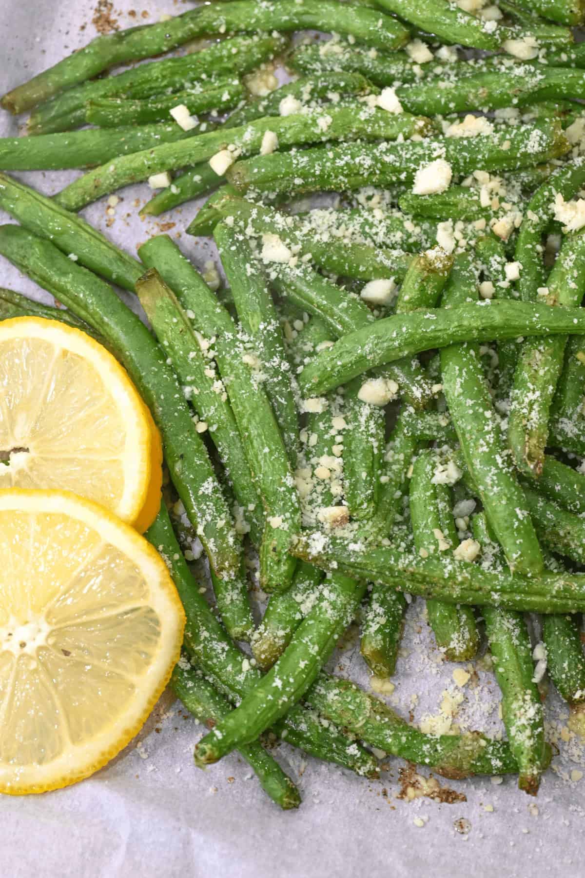 Garlic Parmesan green beans topped with grated cheese sit on a baking sheet, accompanied by two lemon slices for a zesty finish.