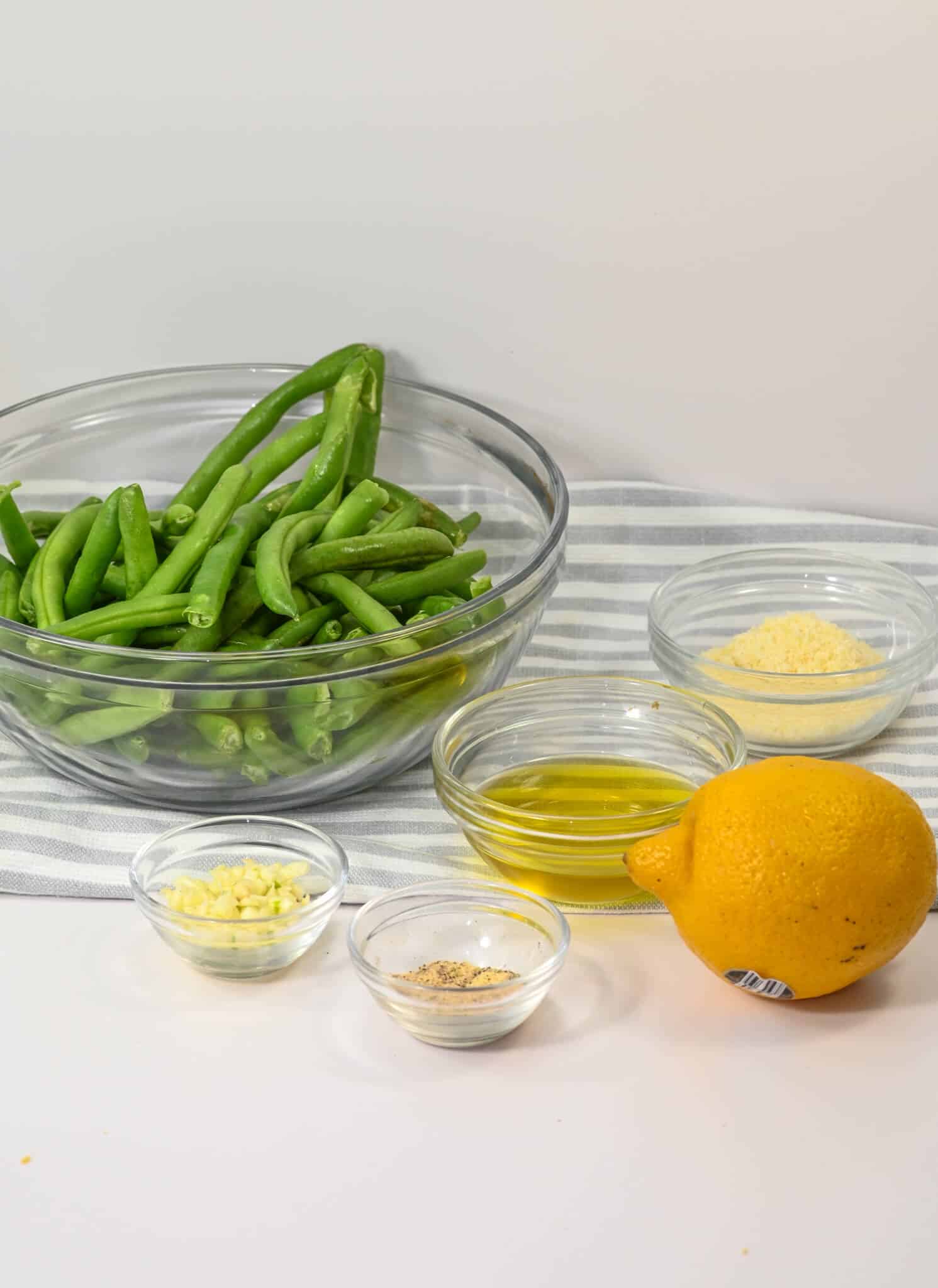 A glass bowl filled with the ingredients sits on a striped cloth, surrounded by lemon, bowls of olive oil, minced garlic, and seasoning.