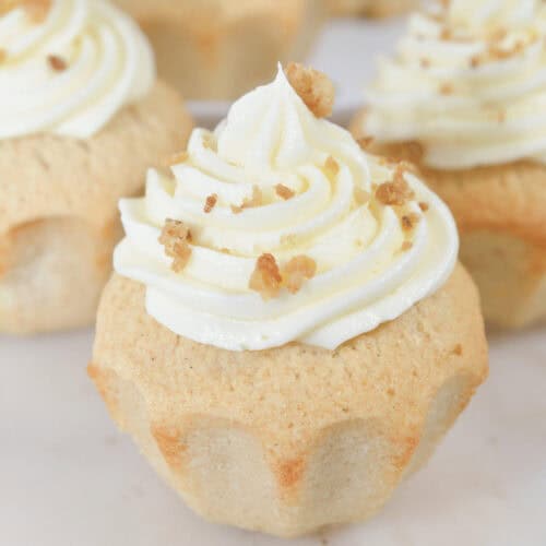 Close-up of four cinnamon cupcakes topped with swirled white frosting and sprinkled with small bits of nuts, arranged on a white marble surface.