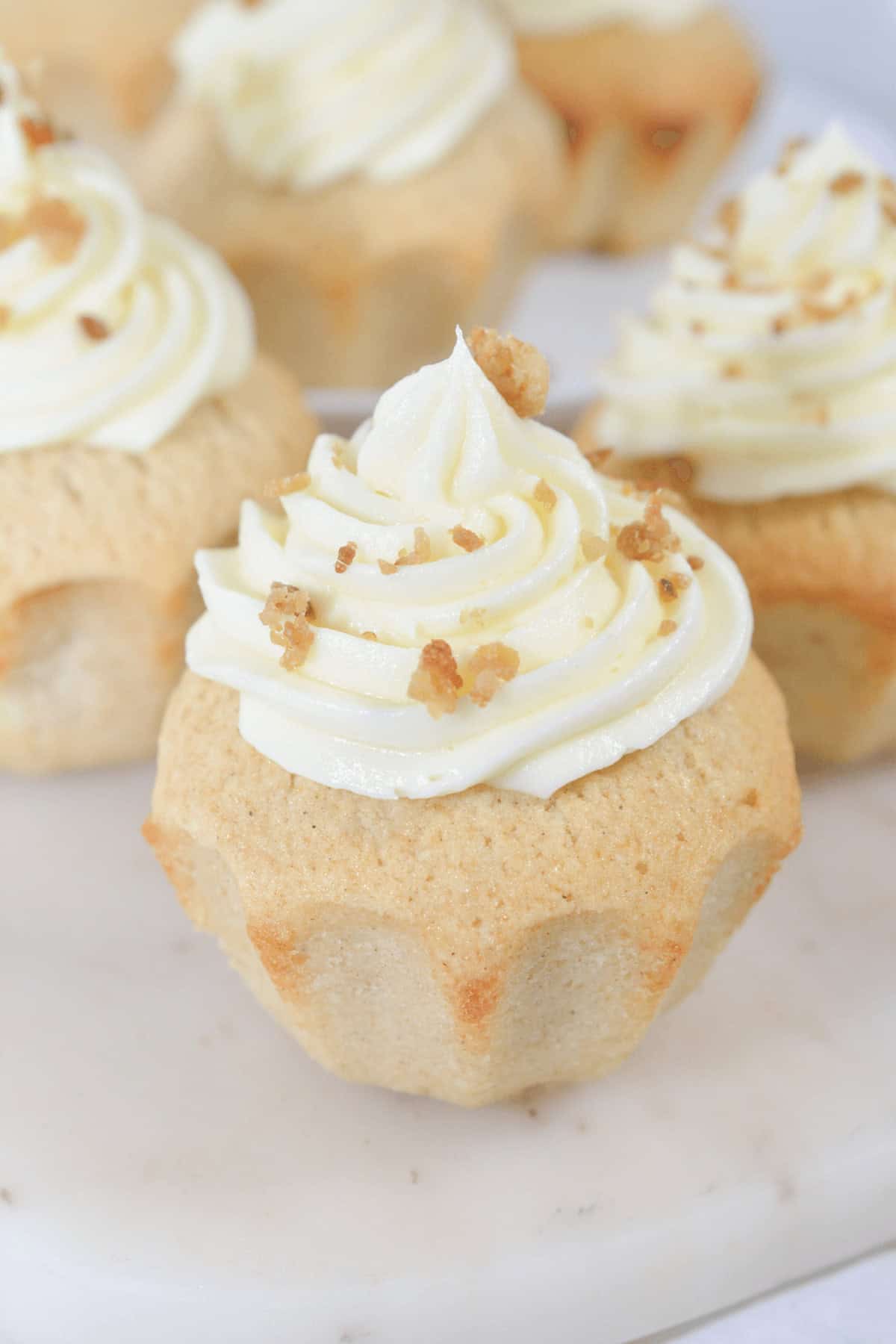 Close-up of four cinnamon cupcakes topped with swirled white frosting and sprinkled with small bits of nuts, arranged on a white marble surface.