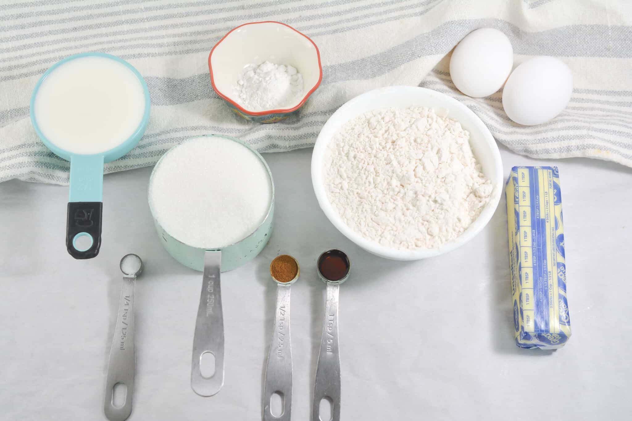 Ingredients laid out on a surface include milk, eggs, flour, butter, sugar, baking powder, vanilla extract, and cinnamon for delicious cinnamon cupcakes, with measuring spoons and cups placed alongside.