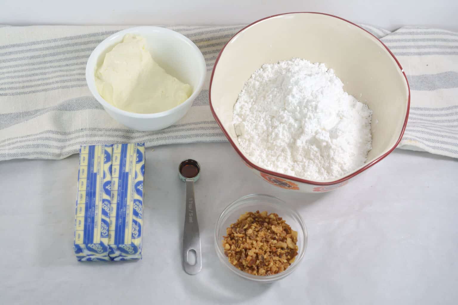 Assorted baking ingredients on a surface: a bowl of cream cheese, powdered sugar in a larger bowl, a stick of butter, a teaspoon of vanilla extract, and a small bowl of chopped nuts—perfect for making cinnamon cupcakes.