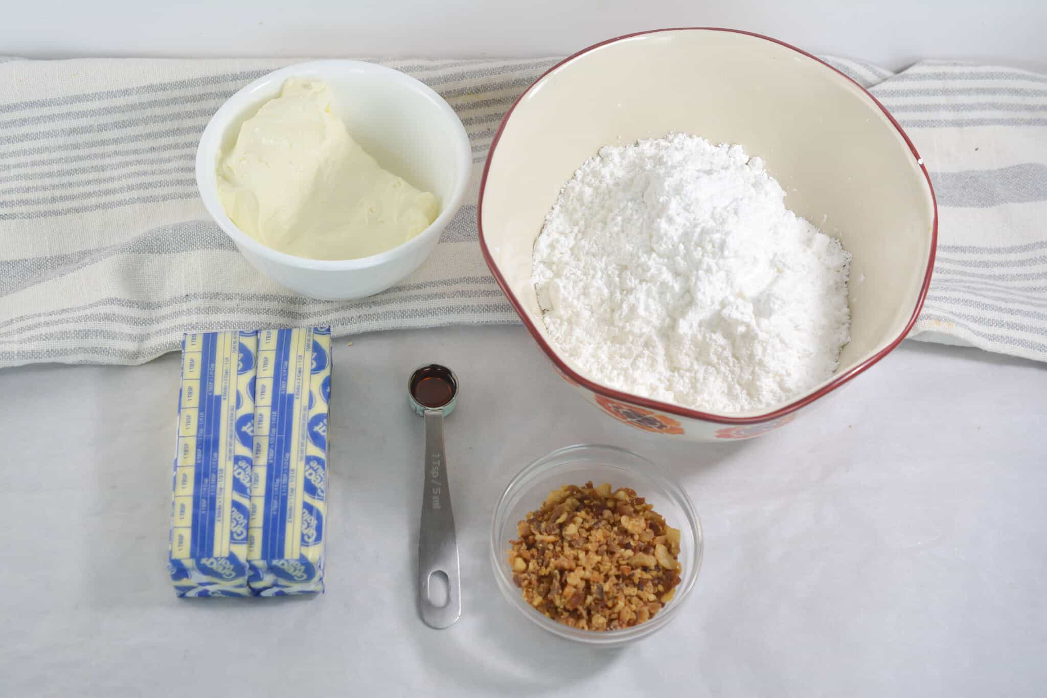 Assorted baking ingredients on a surface: a bowl of cream cheese, powdered sugar in a larger bowl, a stick of butter, a teaspoon of vanilla extract, and a small bowl of chopped nuts—perfect for making cinnamon cupcakes.