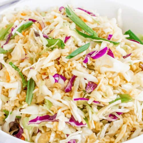 A bowl of coleslaw featuring shredded cabbage, carrots, green onions, and crispy noodles reminiscent of a Ramen Noodle Salad Recipe. Two forks are visible in the background on a white surface.