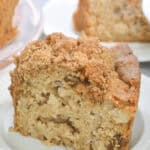 A slice of fresh apple cake with a delicious crumb topping sits invitingly on a white plate, while more cake tempts in the background.