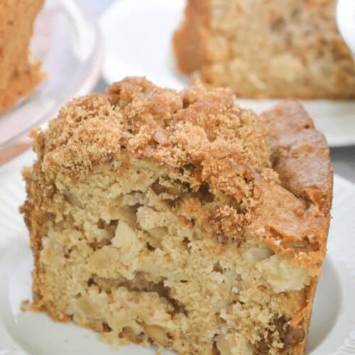 A slice of fresh apple cake with a delicious crumb topping sits invitingly on a white plate, while more cake tempts in the background.
