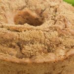 A round bundt cake topped with a crumbly brown layer sits invitingly on a plate, perfectly complemented by the hint of a fresh apple partially visible in the background.