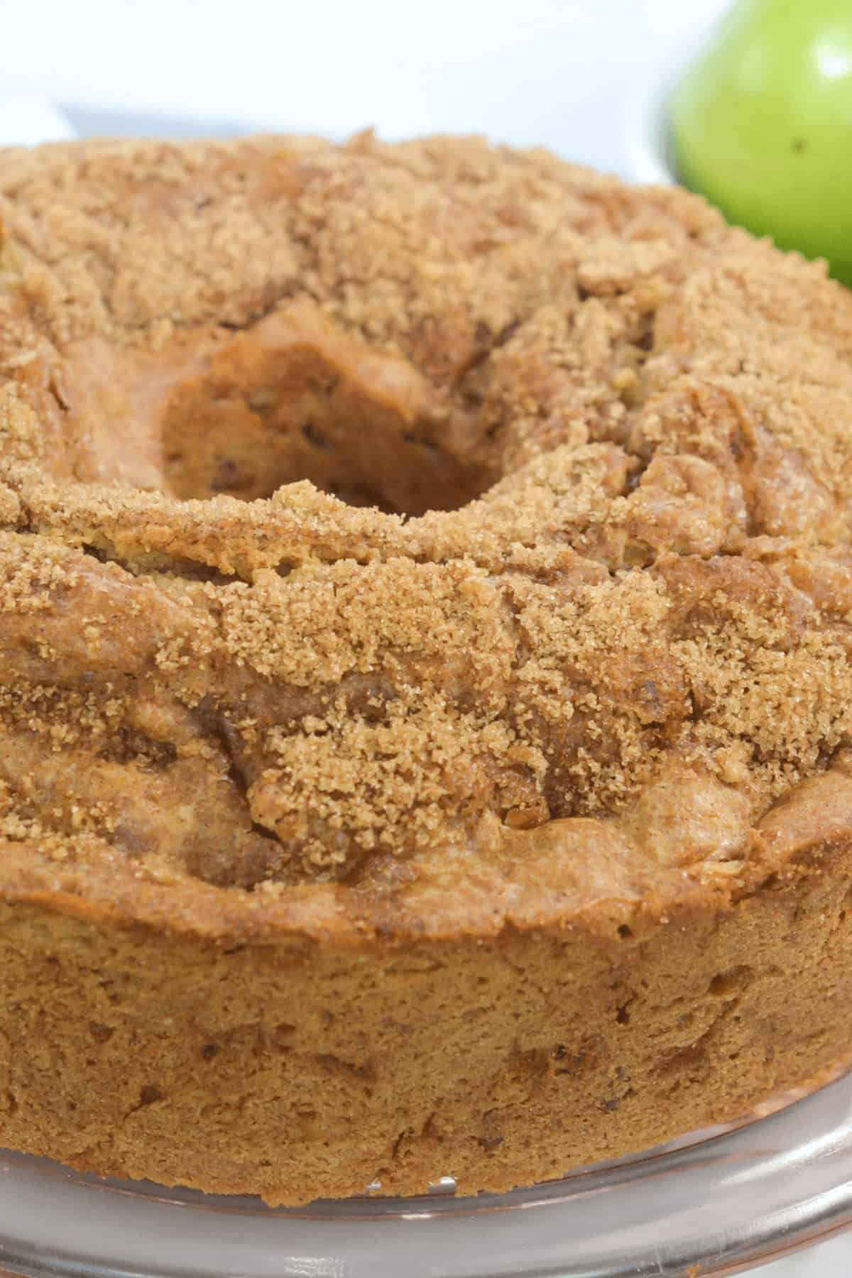 A round bundt cake topped with a crumbly brown layer sits invitingly on a plate, perfectly complemented by the hint of a fresh apple partially visible in the background.
