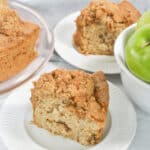 A slice of fresh apple cake on a white plate sits next to a bowl of green apples and a partially visible cake on a platter.