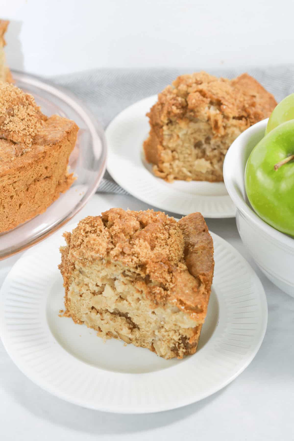 A slice of fresh apple cake on a white plate sits next to a bowl of green apples and a partially visible cake on a platter.