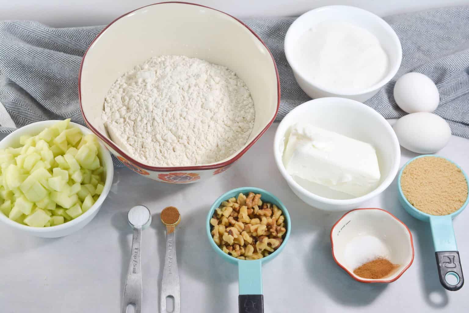 Baking ingredients for the dessert displayed: flour, sugar, eggs, chopped apples, walnuts, cream cheese, brown sugar, cinnamon, nutmeg, and measuring spoons on a table with a grey cloth.