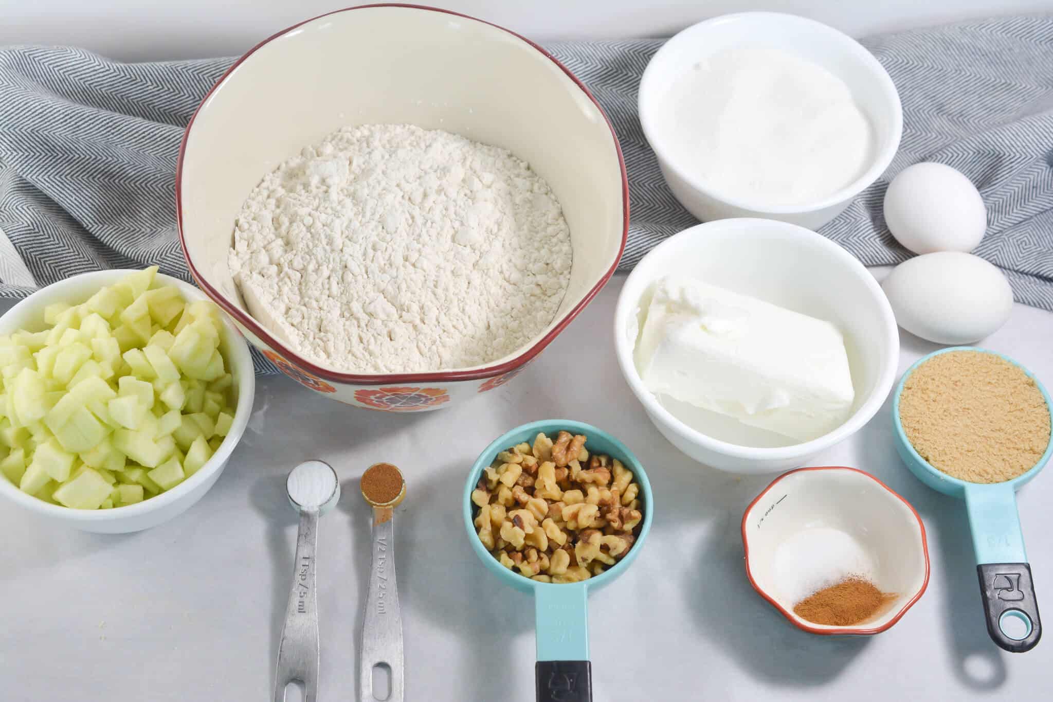 Baking ingredients for the dessert displayed: flour, sugar, eggs, chopped apples, walnuts, cream cheese, brown sugar, cinnamon, nutmeg, and measuring spoons on a table with a grey cloth.