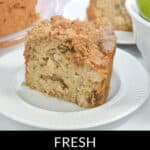 A slice of fresh apple cake with a crumbly top rests on a white plate, sitting beside the whole cake and a bowl of green apples.