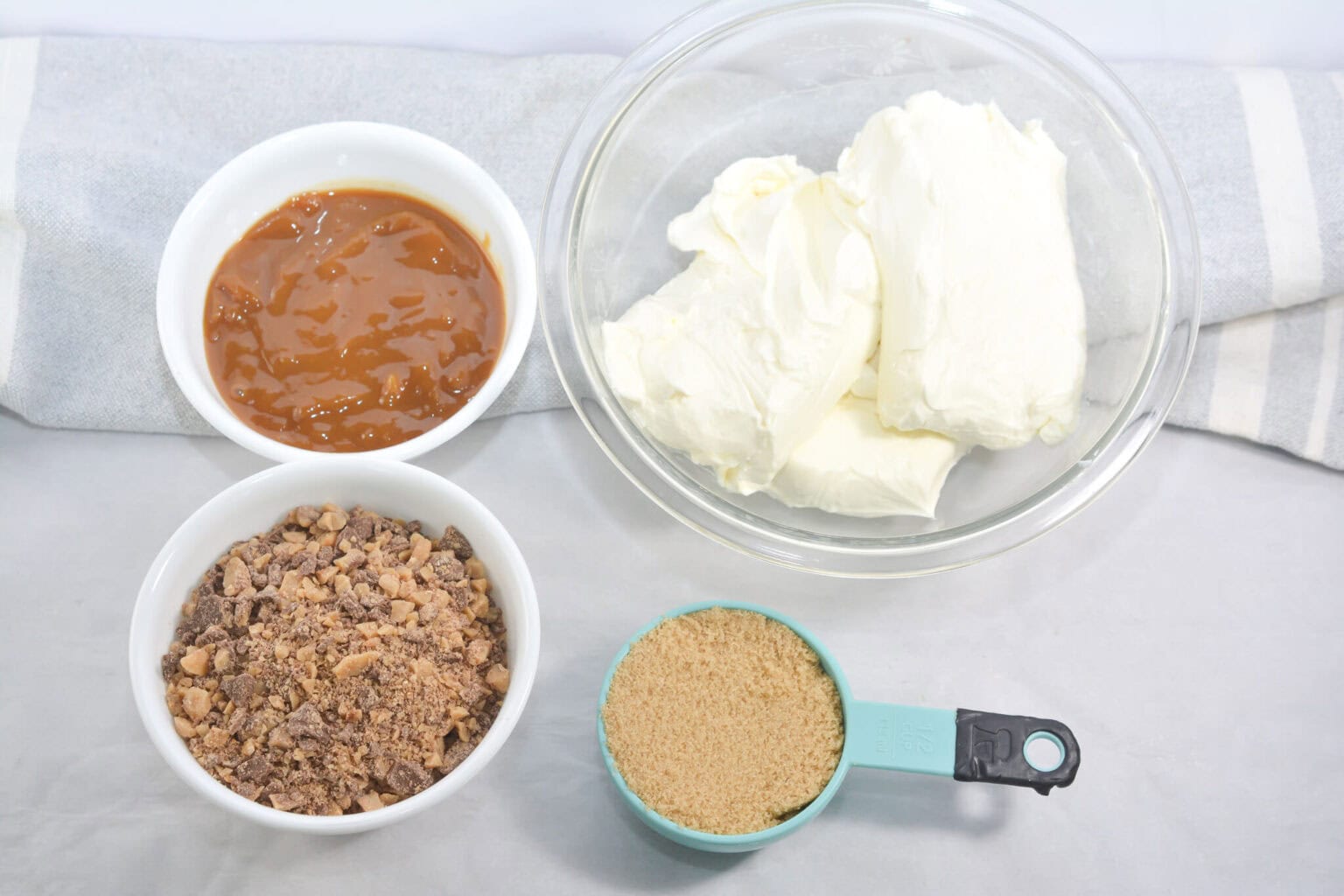Ingredients for a delicious snack are laid out in bowls on a kitchen towel, including whipped cream, caramel sauce, crushed toffee bits, and brown sugar.