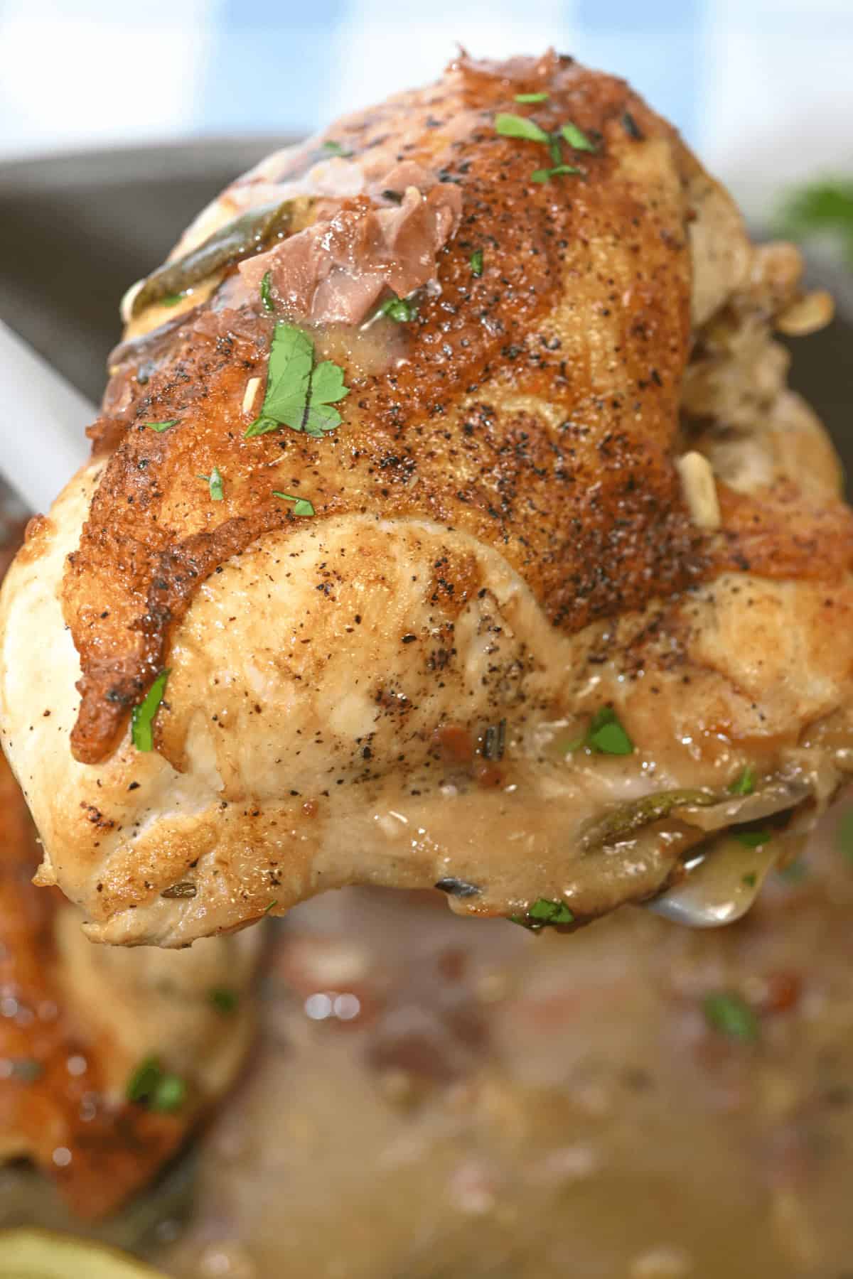 A cooked piece of Herbed Lemon Chicken with a crispy skin is held up with tongs, showcasing the perfect blend of herbs and zesty lemon sauce.