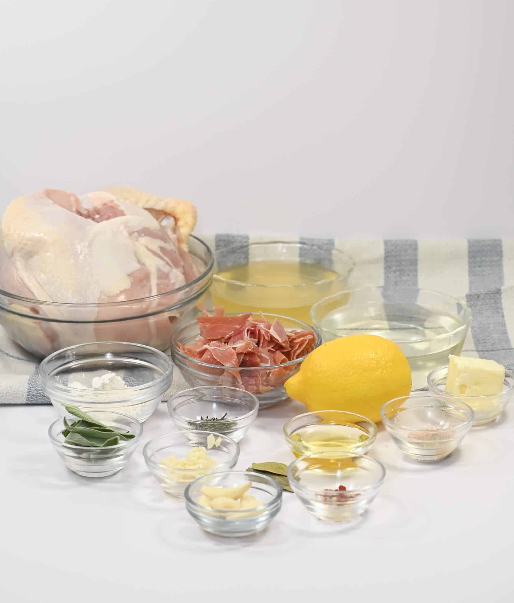 A raw Herbed Lemon Chicken in a bowl is surrounded by small glass bowls containing ingredients like lemon, prosciutto, butter, and various seasonings on a striped cloth background.
