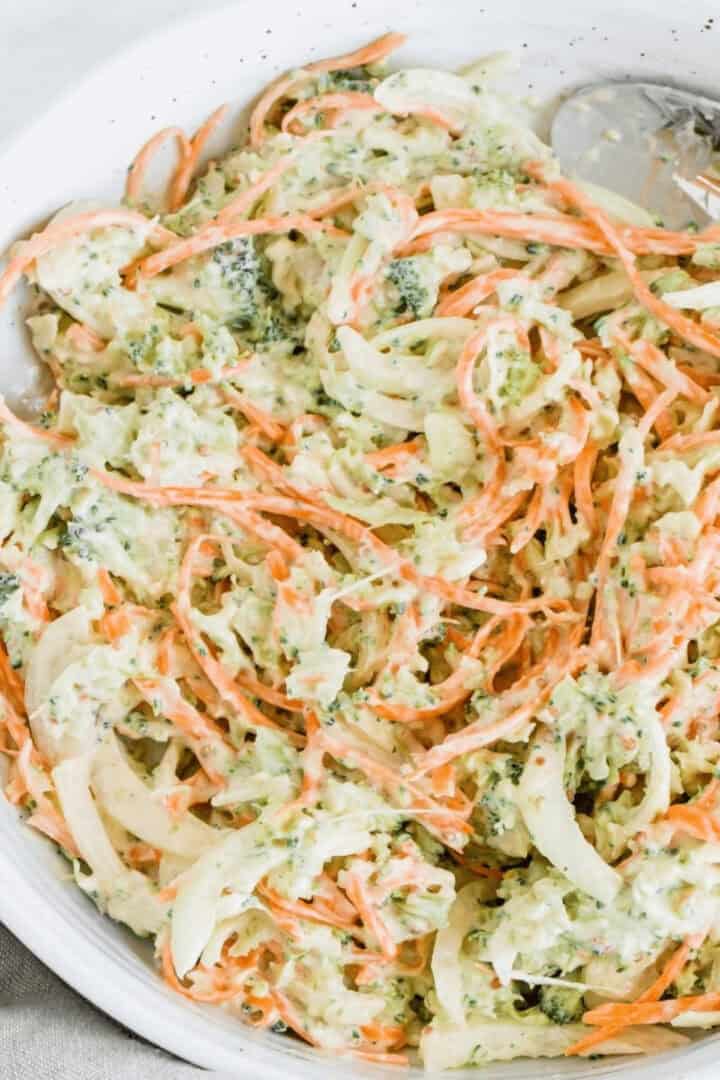 A bowl of creamy broccoli coleslaw with shredded carrots, cabbage, and a hint of broccoli. A serving spoon is on the side.