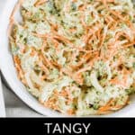 A bowl of broccoli coleslaw with shredded carrots and onions, dressed in a creamy sauce, with a spoon on the side. Text overlay reads "Tangy Broccoli Coleslaw.