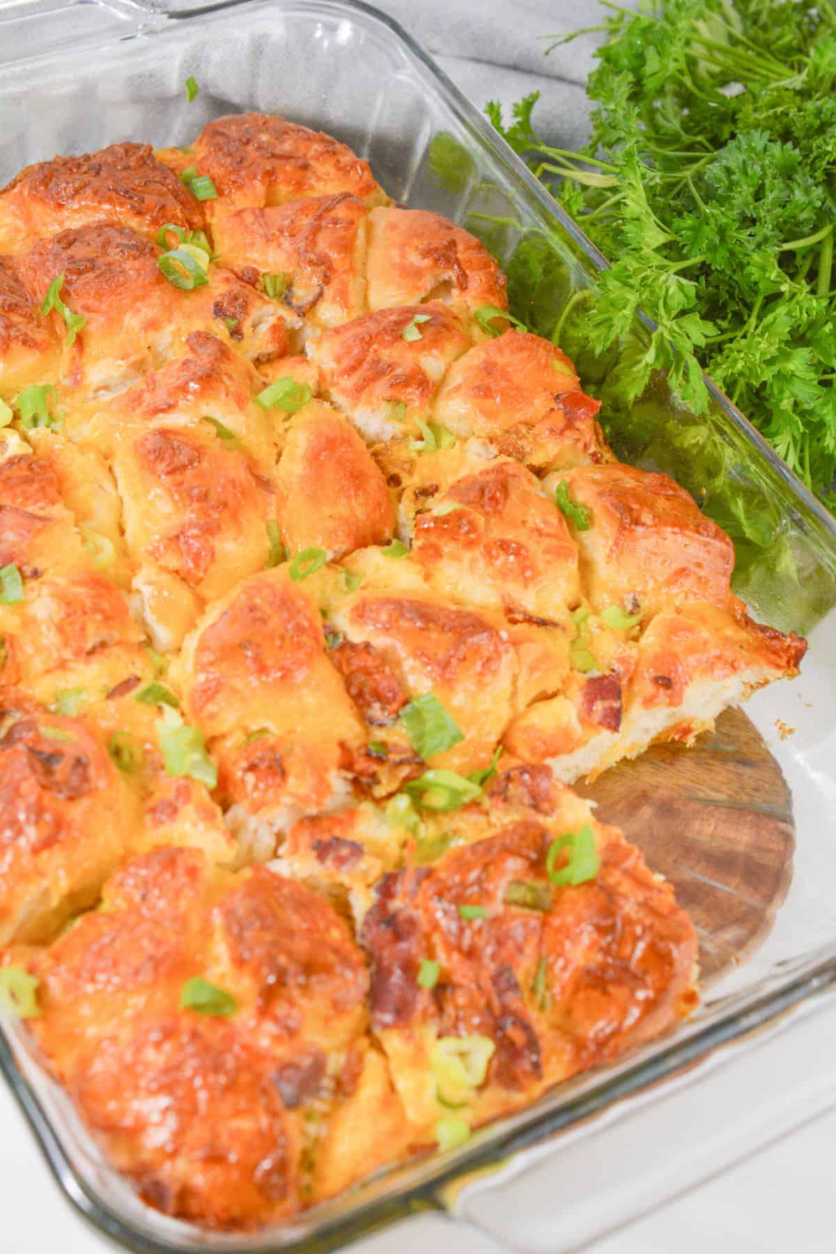 A glass baking dish filled with golden-brown Biscuit Breakfast Casserole rolls, topped with chopped green herbs, sits next to a bunch of fresh parsley.