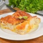 A slice of Biscuit Breakfast Casserole with a golden crust sits on a white plate, garnished with chopped green onions, with herbs and utensils in the background.
