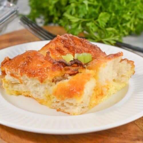 A slice of Biscuit Breakfast Casserole with a golden crust sits on a white plate, garnished with chopped green onions, with herbs and utensils in the background.