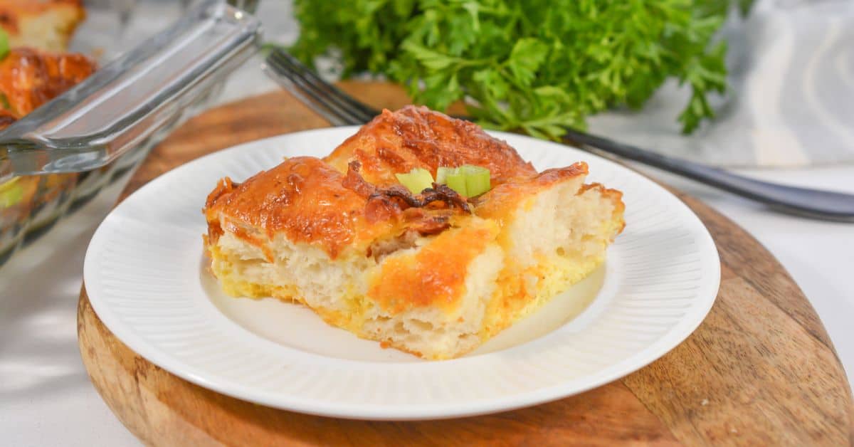 A slice of Biscuit Breakfast Casserole with a golden crust sits on a white plate, garnished with chopped green onions, with herbs and utensils in the background.