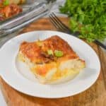 A slice of Biscuit Breakfast Casserole topped with chopped green onions sits on a white plate, next to a glass baking dish and fresh parsley on a wooden board.