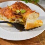 A slice of Biscuit Breakfast Casserole topped with green onions sits on a white plate, with a fork holding a bite-sized piece.
