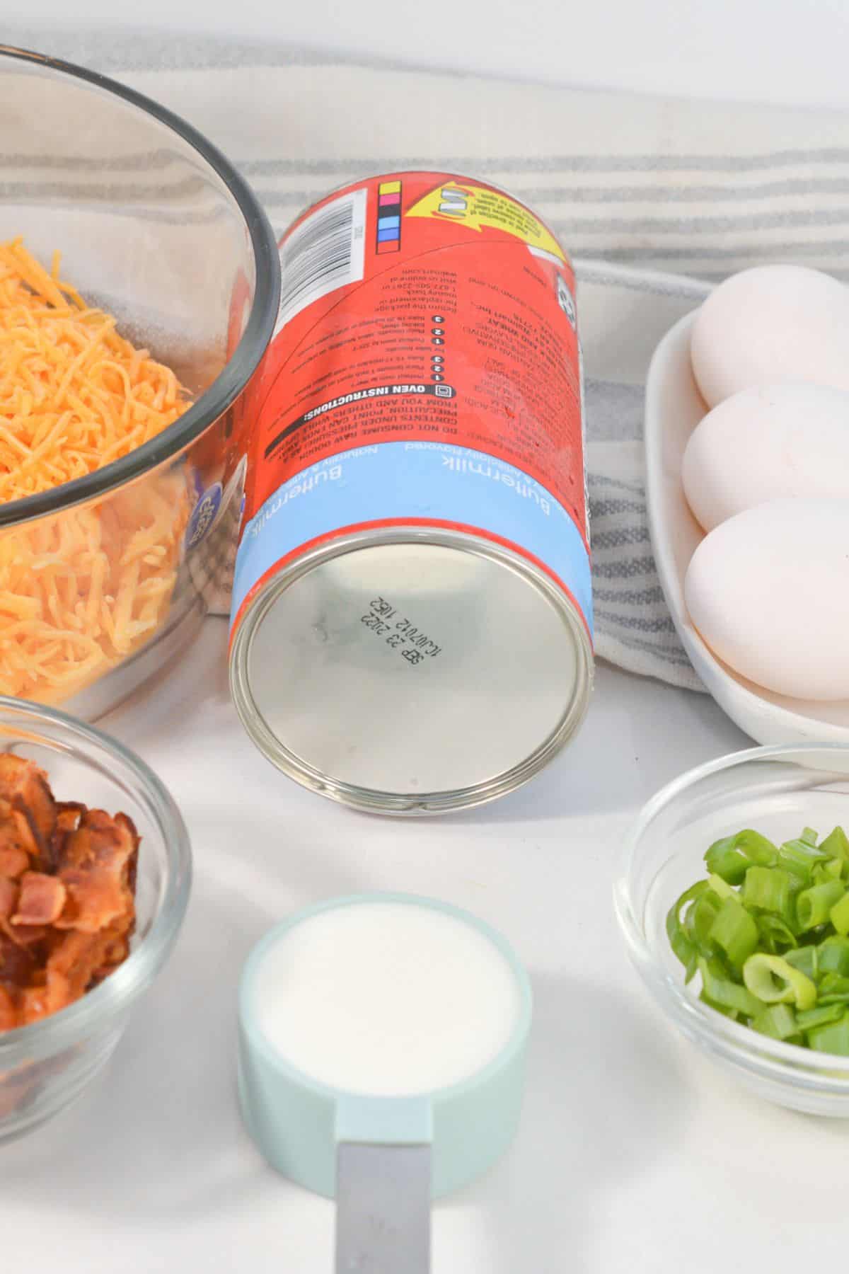 Ingredients  laid out on a white surface, including shredded cheese, canned buttermilk biscuits, eggs, cooked bacon, green onions, and a measuring cup of milk.