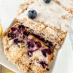 A lemon blueberry loaf, topped with white glaze and whole blueberries, sits beautifully sliced on a white plate.