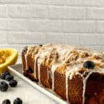 A rectangular lemon blueberry loaf with white glaze on top is placed on a white plate. The cake is garnished with whole blueberries and is accompanied by a halved lemon and scattered blueberries.